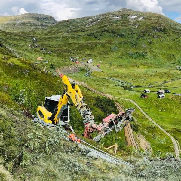 Gravemaskin arbeider på en bratt skråning med grønne åser og hytter i bakgrunnen.