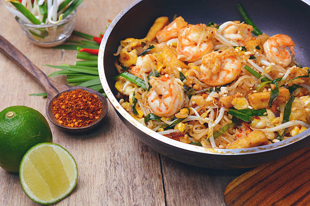 En bolle med pad thai med reker, bønnespirer, lime og krydder på et trebord.