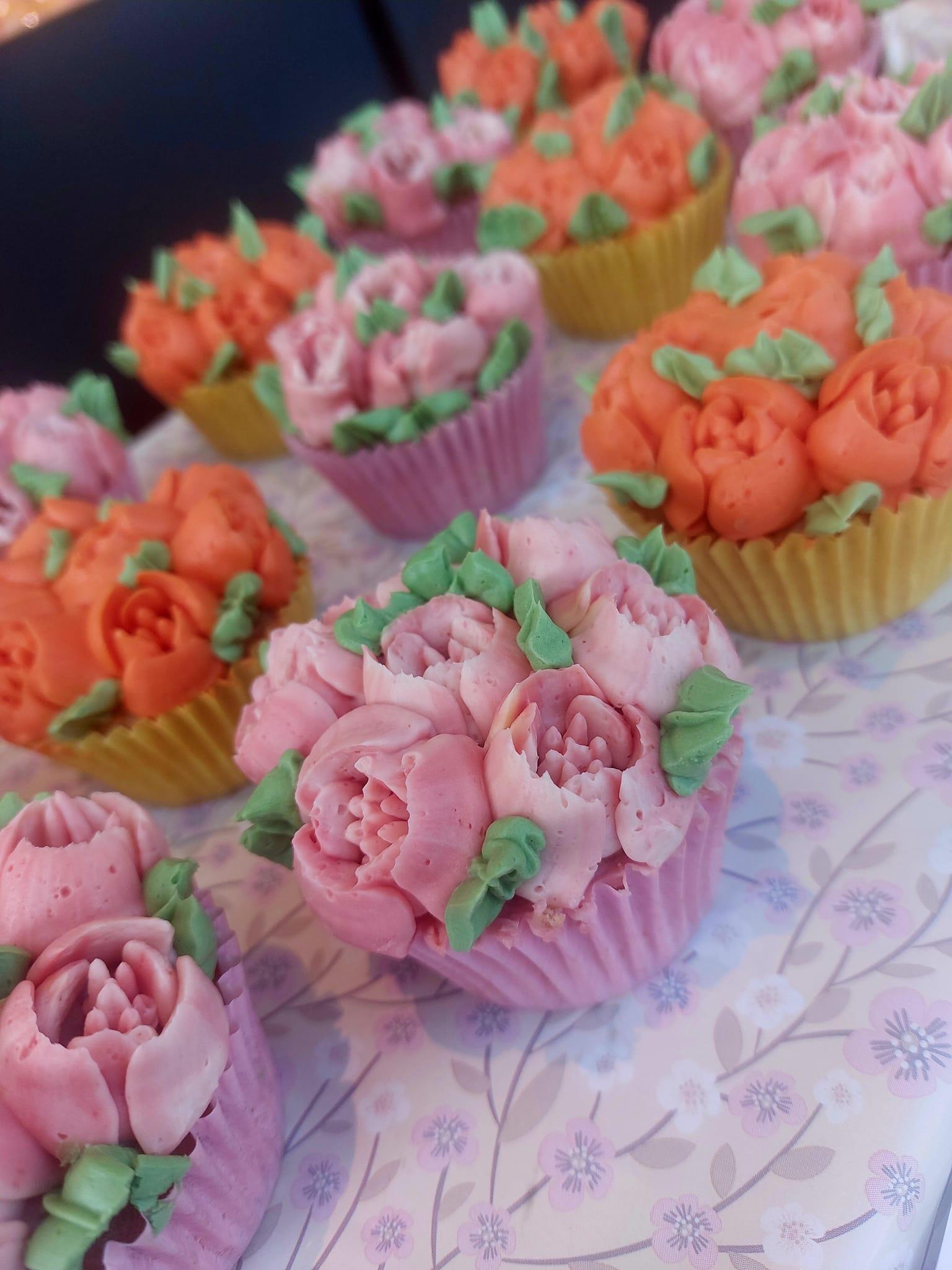 Fargerike cupcakes med blomsterdekorasjoner i rosa og oransje, plassert på et mønstret bord.