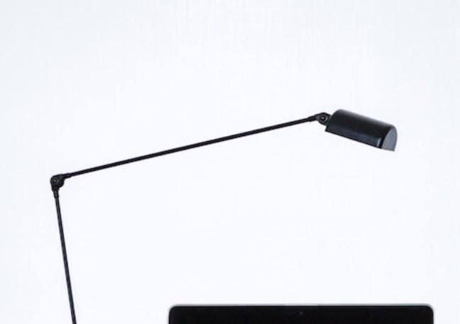 En svart skrivebordslampe med justerbar arm over en laptop på et lyst bord.