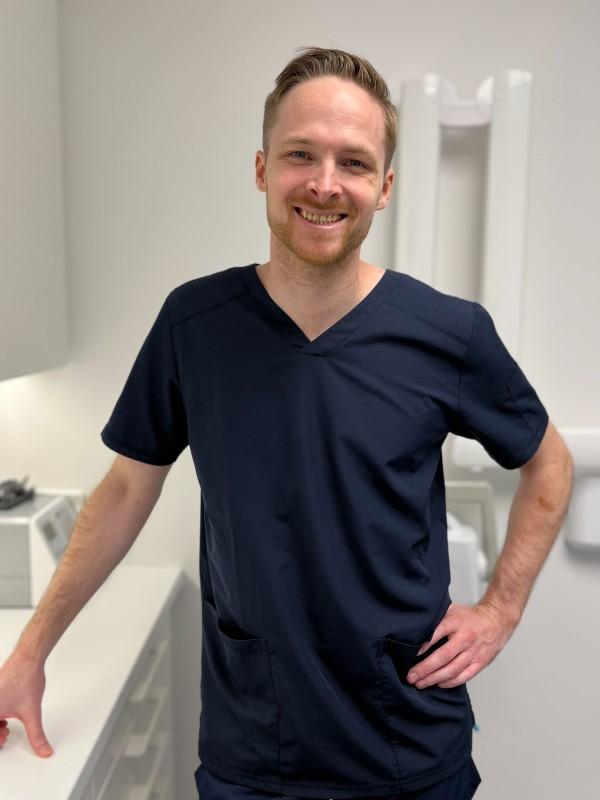En smilende mann i mørkeblå legeuniform står ved en hvit arbeidsbenk i et moderne kontor.