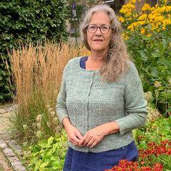 Kvinne med grått hår iført grønn genser står foran blomster i en hage med høye gress og gule blomster.