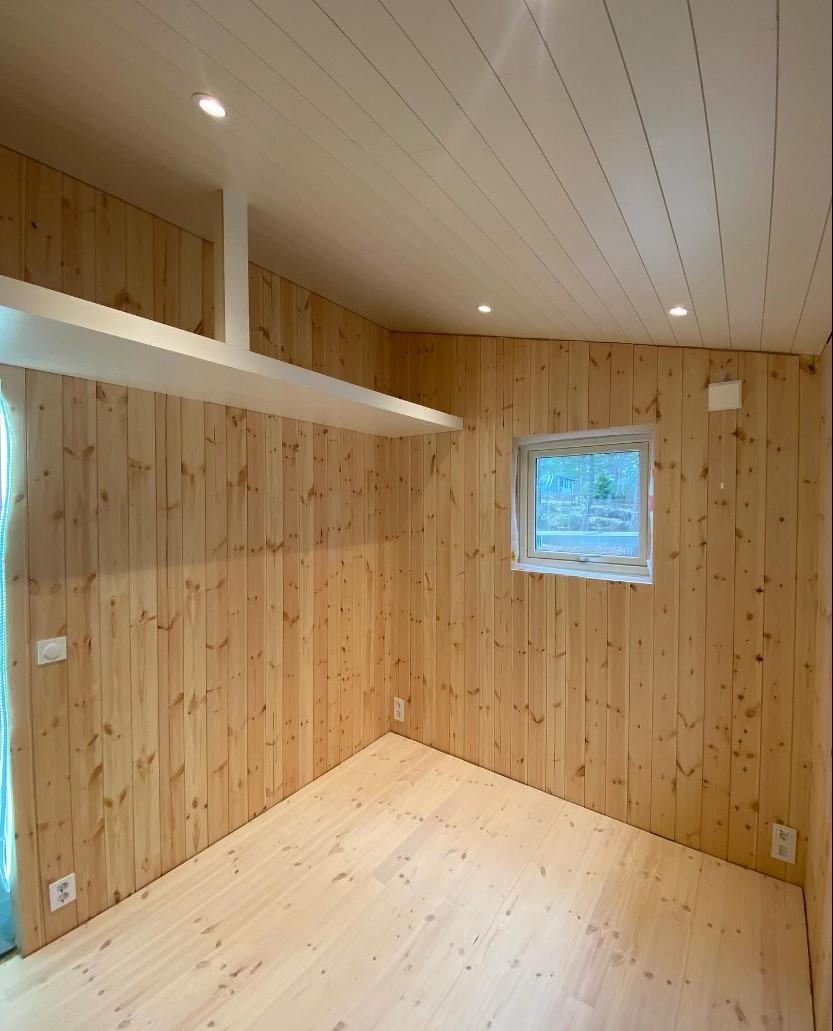 Lyst rom med panelvegger i tre, taklamper og et vindu som slipper inn naturlig lys.