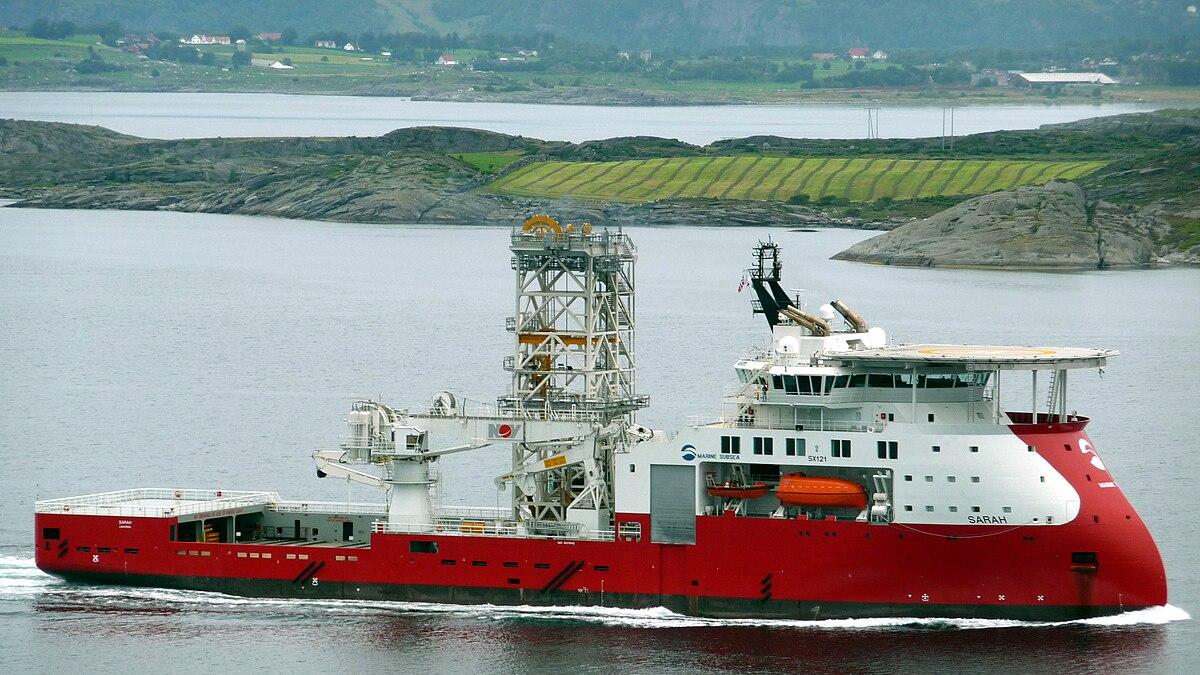 Et rødt offshore-fartøy seiler gjennom vannet med en fjord og grønne åser i bakgrunnen.