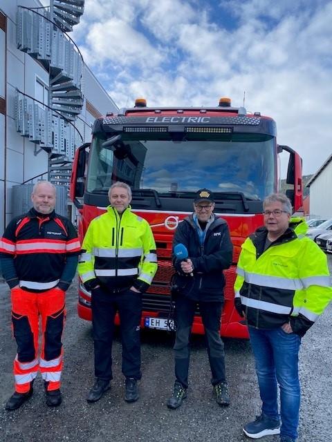 Fire menn står foran en rød elektrisk lastebil, iført refleksjakker og arbeidsklær, med en bygning i bakgrunnen.