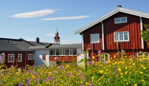 Rødt hus med hvite vinduer omgitt av blomstereng under en blå himmel med skyer.