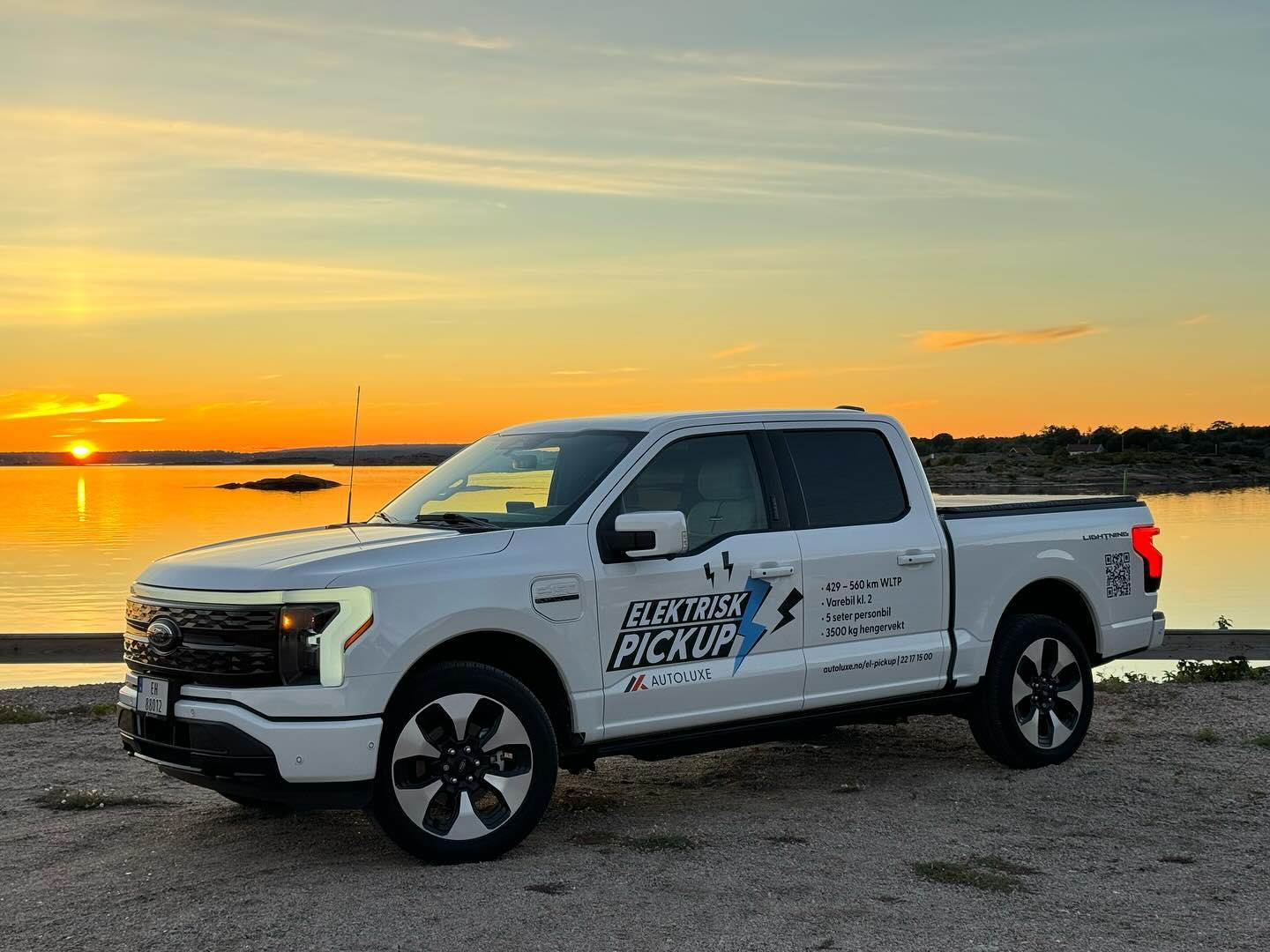 En hvit pickup med logo står ved vannet under en solnedgang. Refleksjoner i vannet skaper en vakker atmosfære.