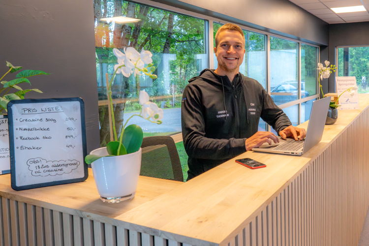 En smilende mann sitter ved resepsjonen med en laptop, omgitt av planter og informasjonstavle.