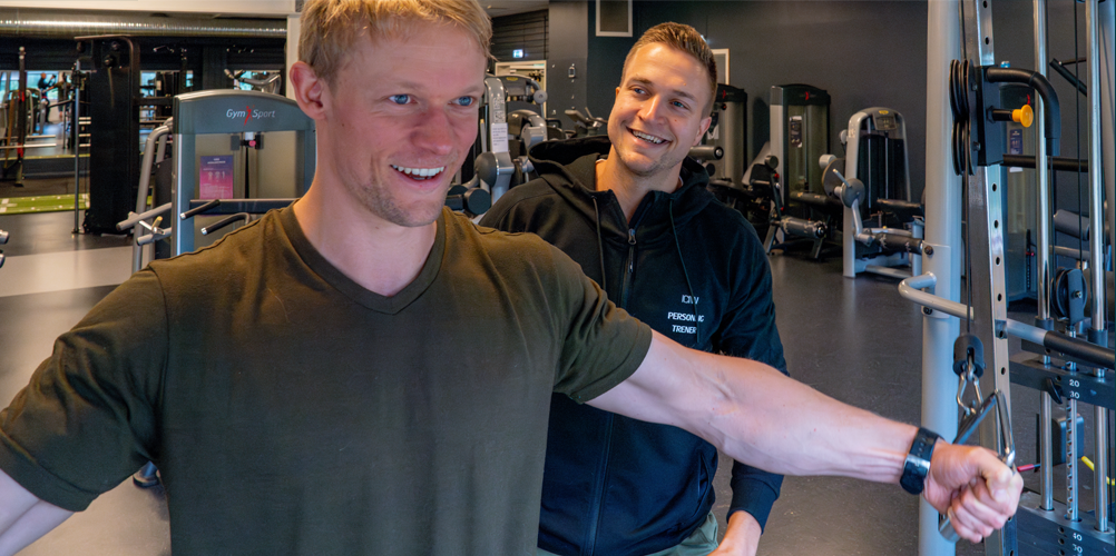 To personer i et treningsstudio, en mann trener med vekter mens en annen smiler og gir veiledning.