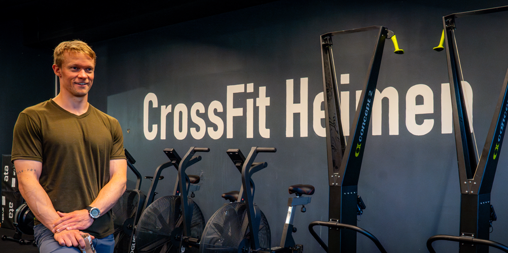 En mann står foran en vegg med teksten "CrossFit Heimen" i et treningsstudio med treningsutstyr.
