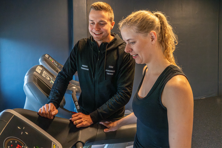 Trener gir veiledning til kvinne på tredemølle i treningsstudio. Begge smiler og ser mot skjermen.