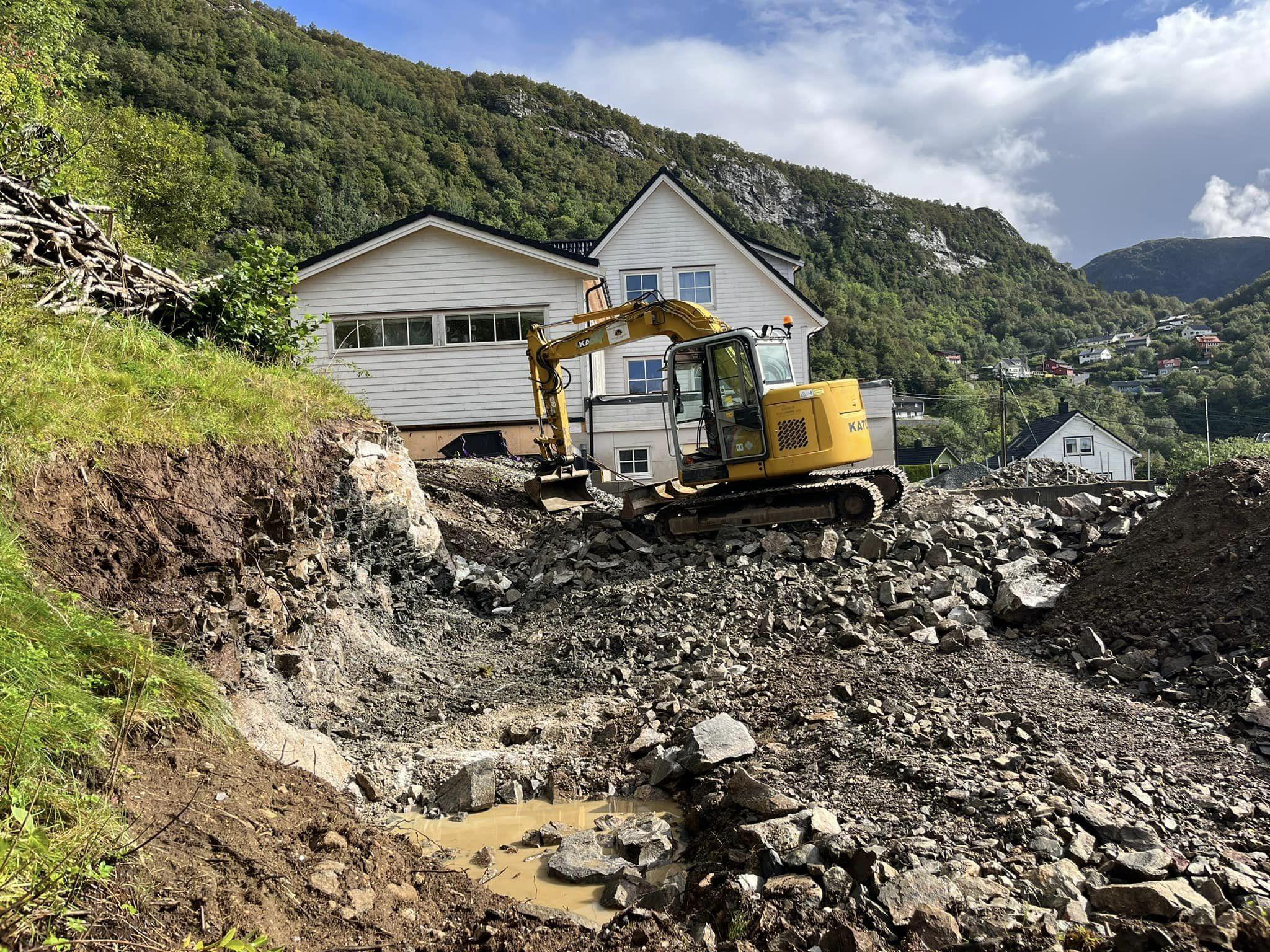 Gravemaskin arbeider på en byggeplass med stein og grus, med hus og fjell i bakgrunnen.