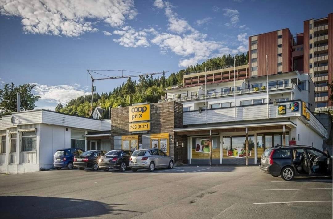 Coop butikk med parkeringsplass og bygninger i bakgrunnen, omgitt av grønt landskap og blå himmel.