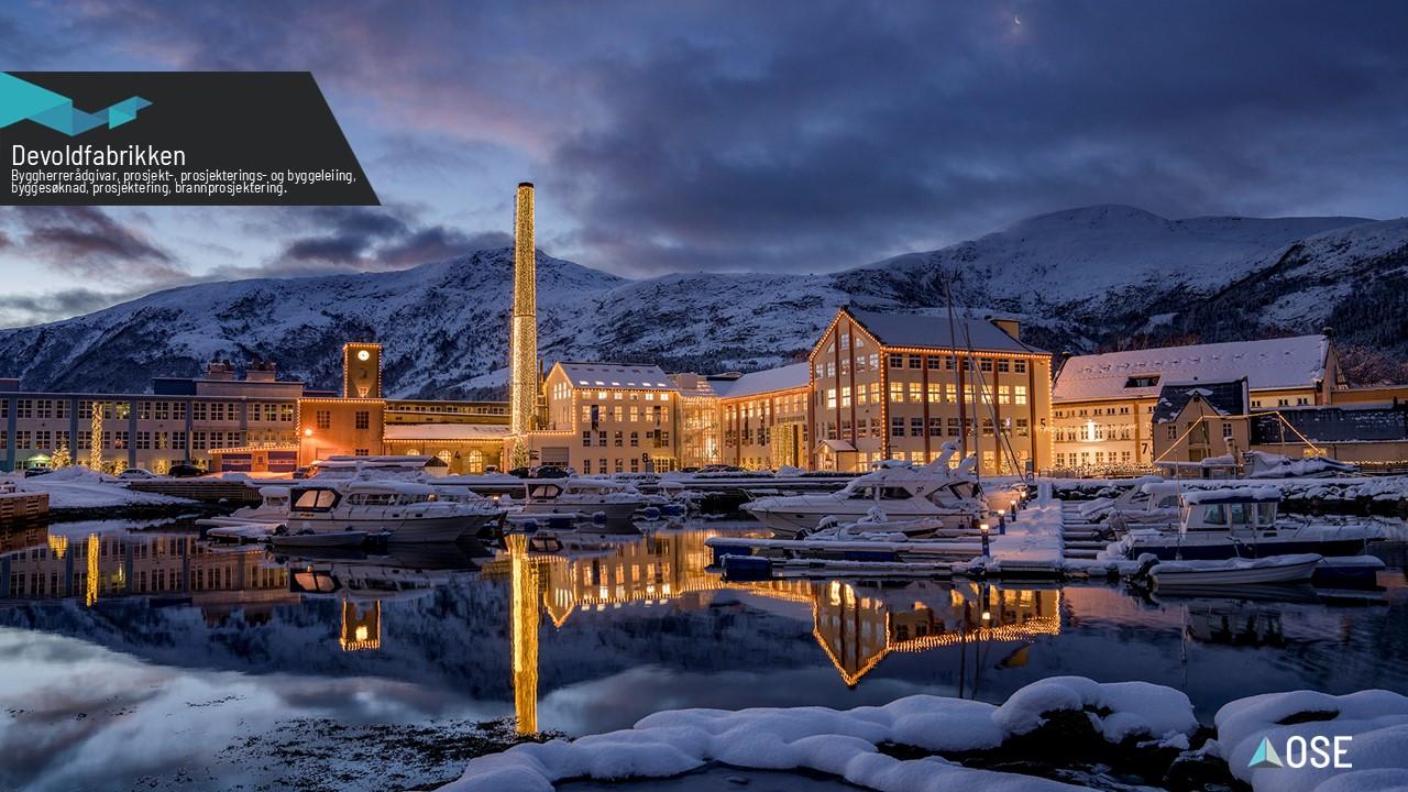 En vakker utsikt over en opplyst bygning ved vannet, med snødekte fjell i bakgrunnen og båter i havnen.