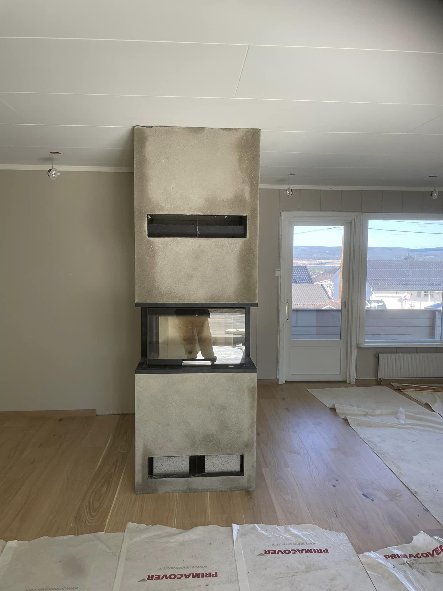 Moderne peis i betong med glassfront, plassert i et rom med lys parkettgulv og store vinduer.
