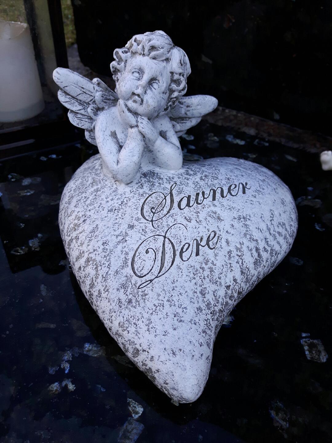 En hvit engel med vinger som sitter på et hjerte med teksten "Savner Dere" på en gravstein.
