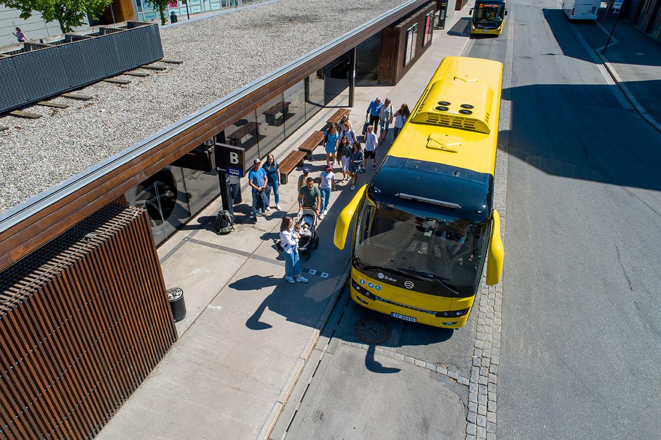 En gul buss står ved busstoppet, mens passasjerer venter og går ombord på en solfylt dag.