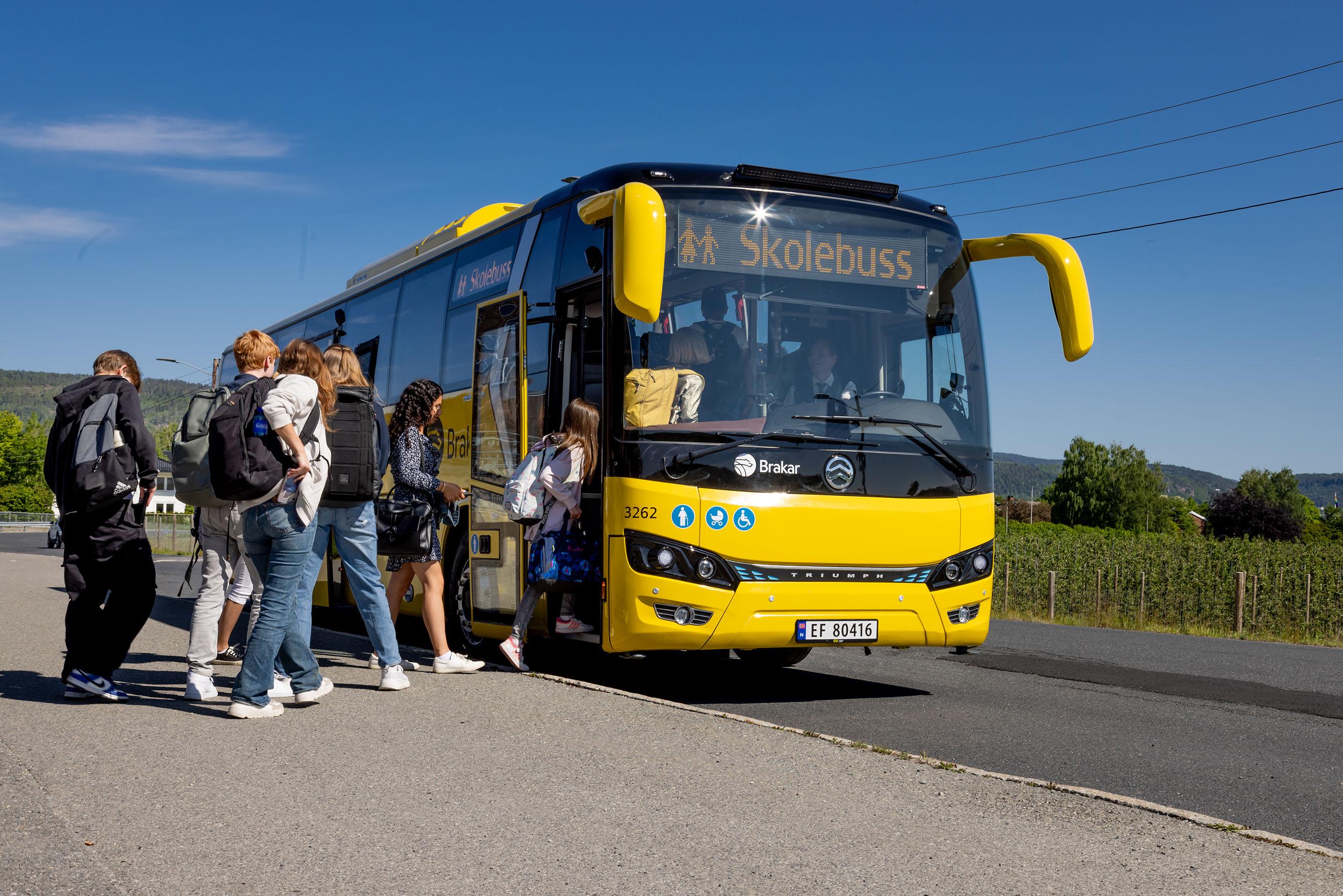 En skolebuss med gule detaljer står parkert ved veien, mens elever går ombord i bussen en solfylt dag.