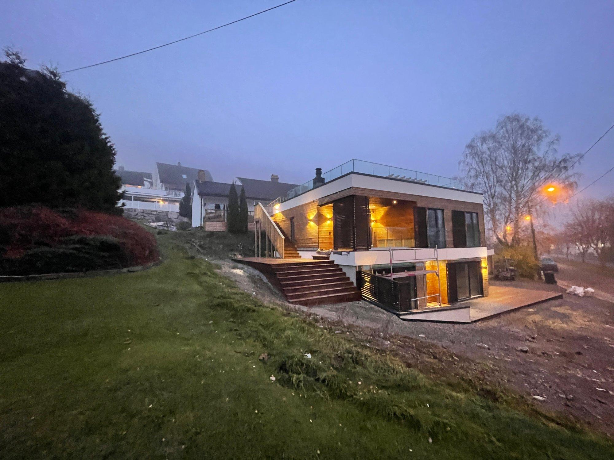 Moderne hus med store vinduer og terrasse, omgitt av grønt landskap i tåke. Belysning gir en koselig atmosfære.