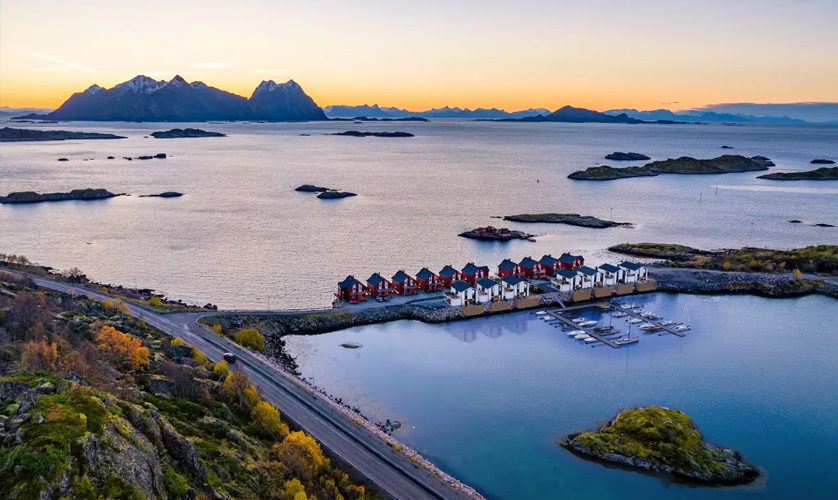 En vakker utsikt over en kystlinje med fargerike hus, båter og fjell i bakgrunnen ved solnedgang.