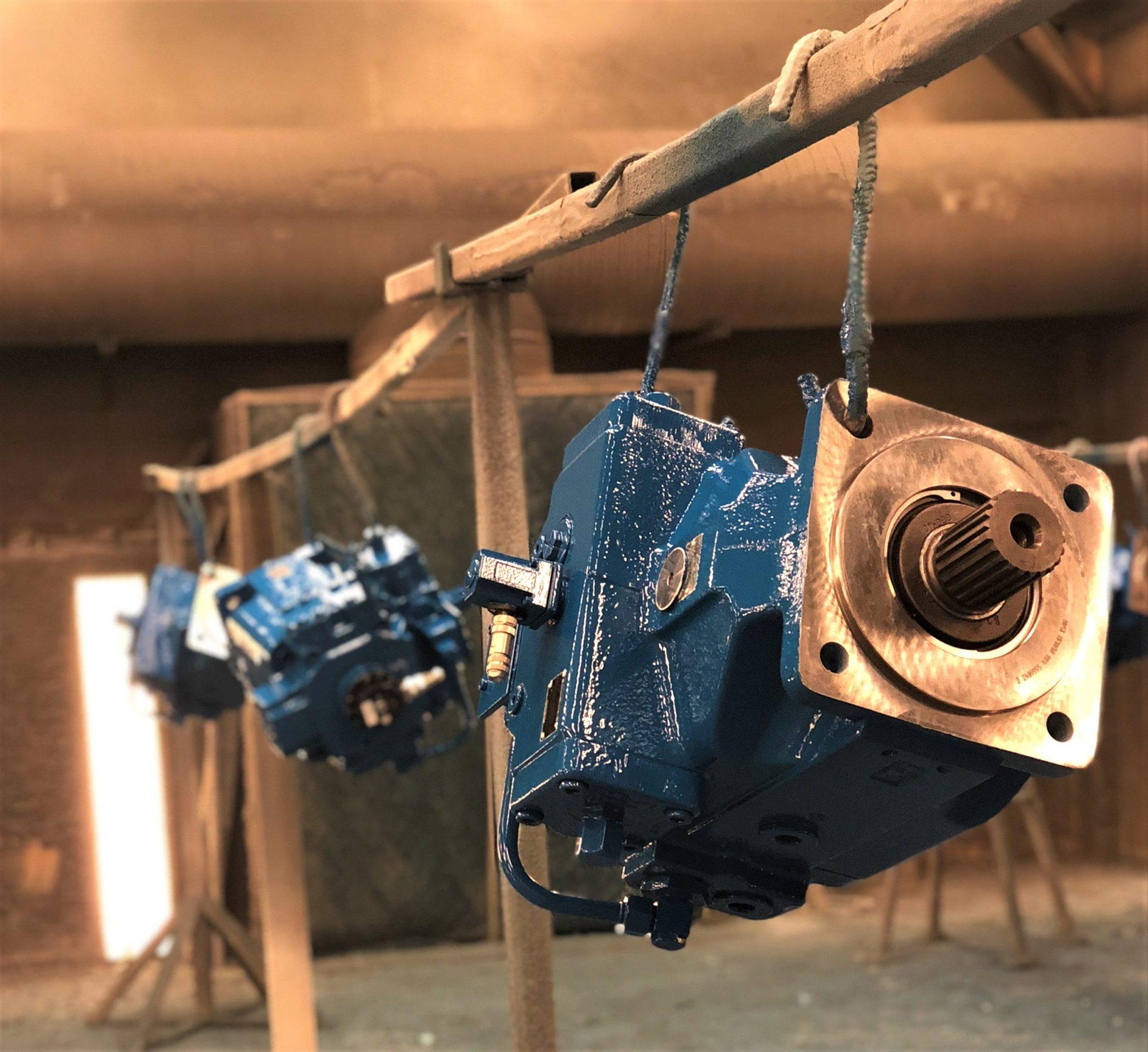 Blå hydraulikkpumper hengende på en stang i et malerom, med fokus på en av pumpene i forgrunnen.