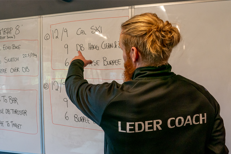En trener peker på en tavle med treningsprogram og øvelser i et treningsstudio.