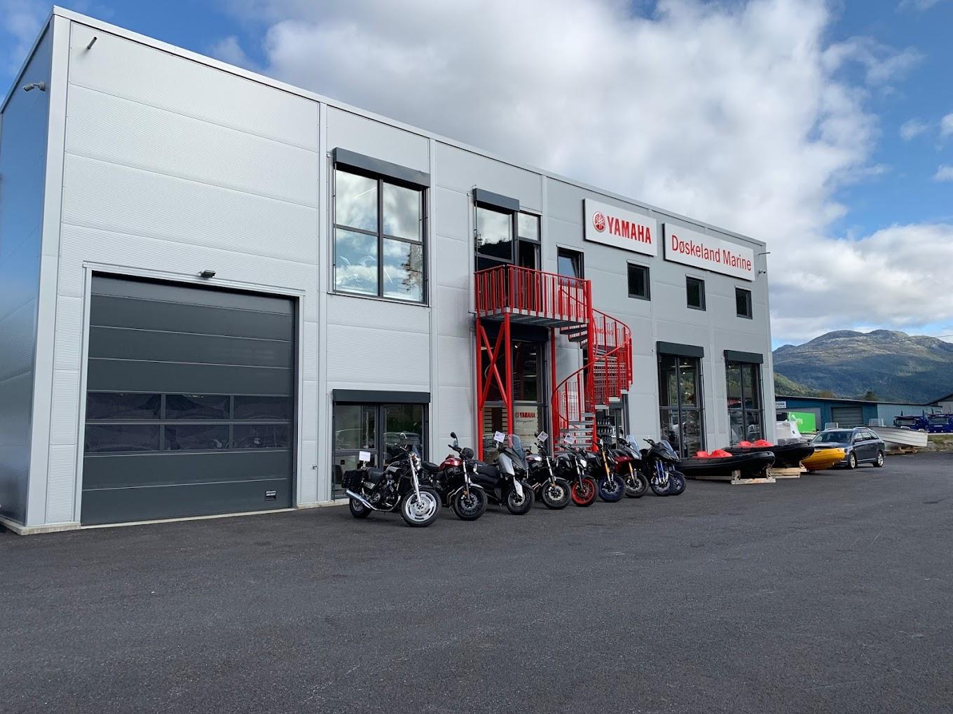 Moderne bygning med rød branntrapp, flere motorsykler og båter parkert foran. Fjell i bakgrunnen.