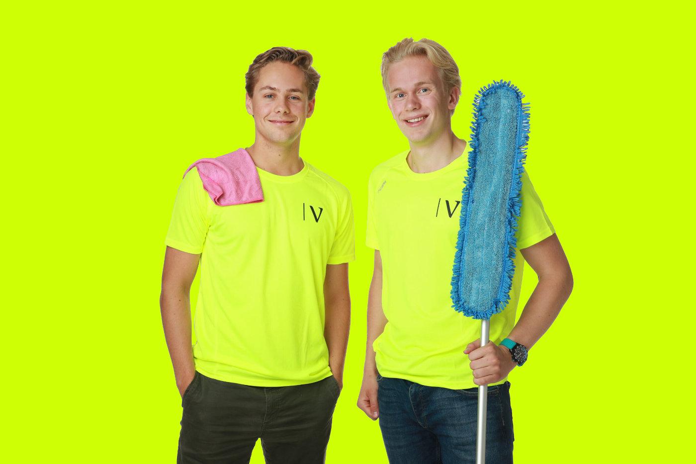 To unge menn i neon gule t-skjorter, en med en klut på skulderen og den andre med en støvkost.