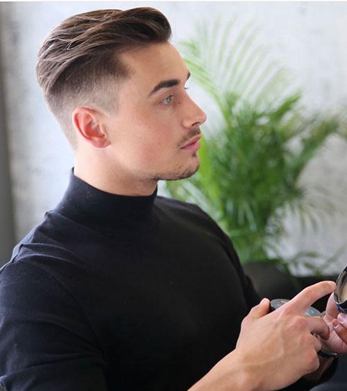 En ung mann med kort hår i svart genser, som ser på en telefon i et moderne rom med planter i bakgrunnen.