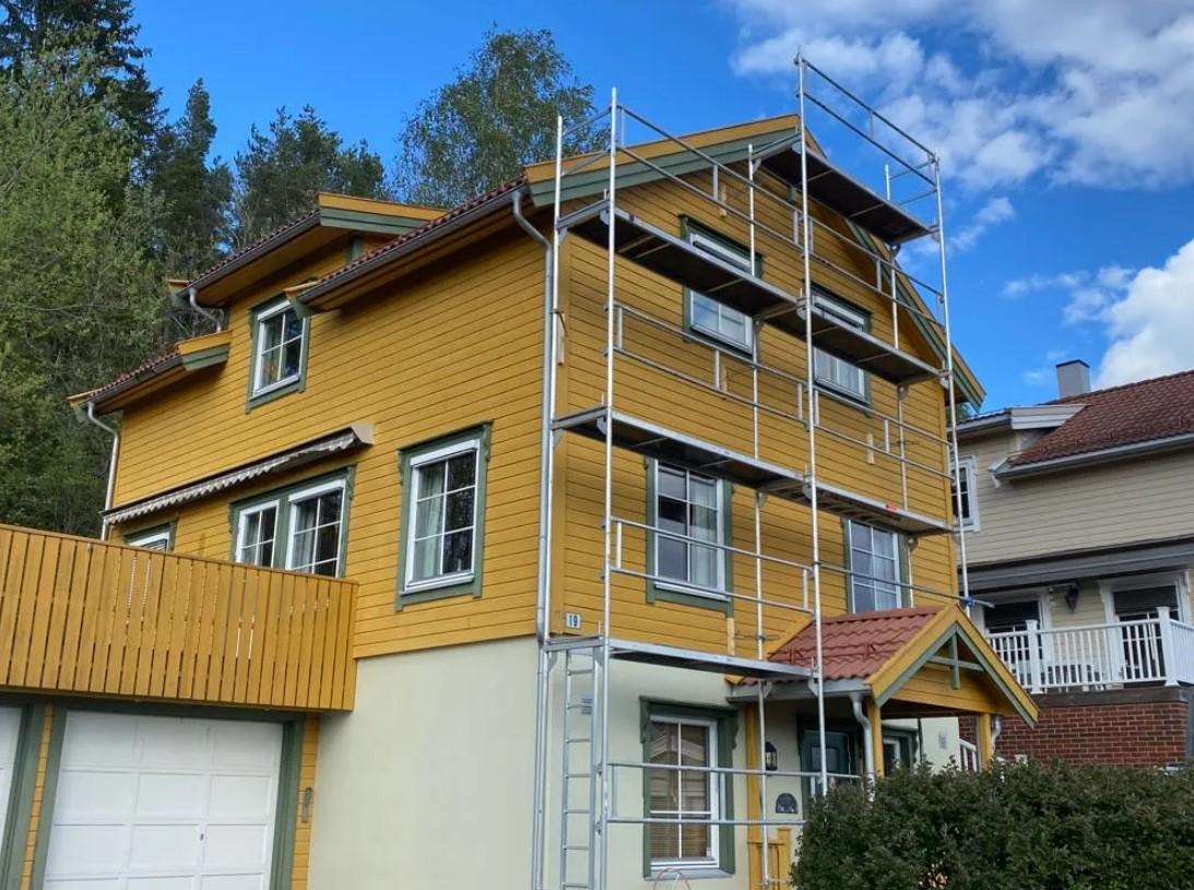 Gult hus med stillas, omgitt av trær og andre bygninger, under oppussing i et boligområde.