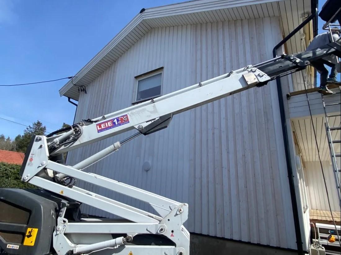 En arbeidsplattform i bruk ved en hvit husvegg, med en person som jobber på en stige.