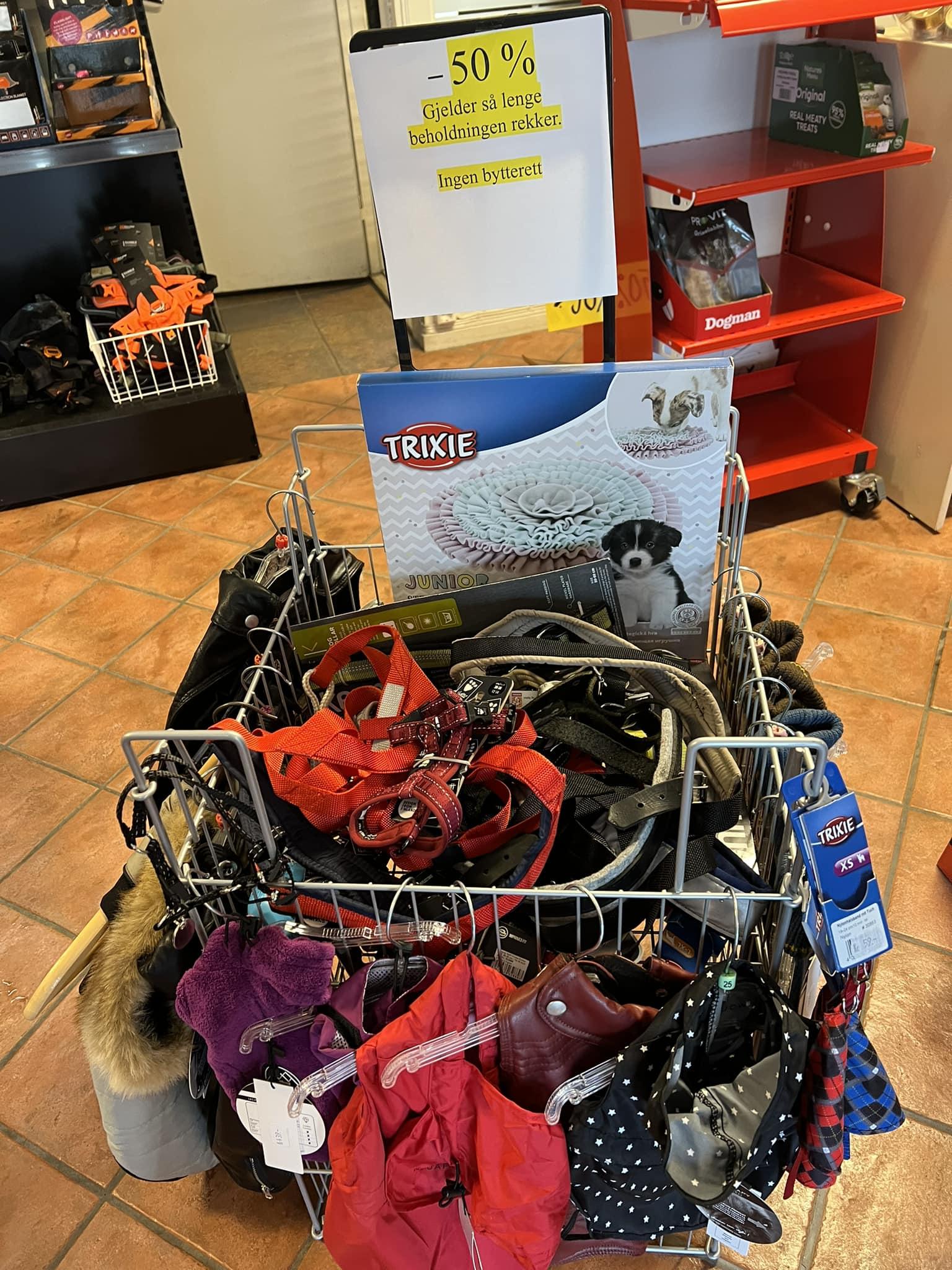 En kurv med hundeutstyr, inkludert bånd, seler og leker, med et skilt som viser 50% rabatt.