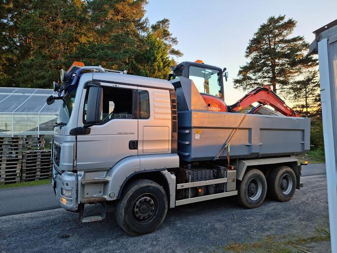 En sølvgrå lastebil med gravemaskin på lasteplanet, parkert ved en skogkledd vei.
