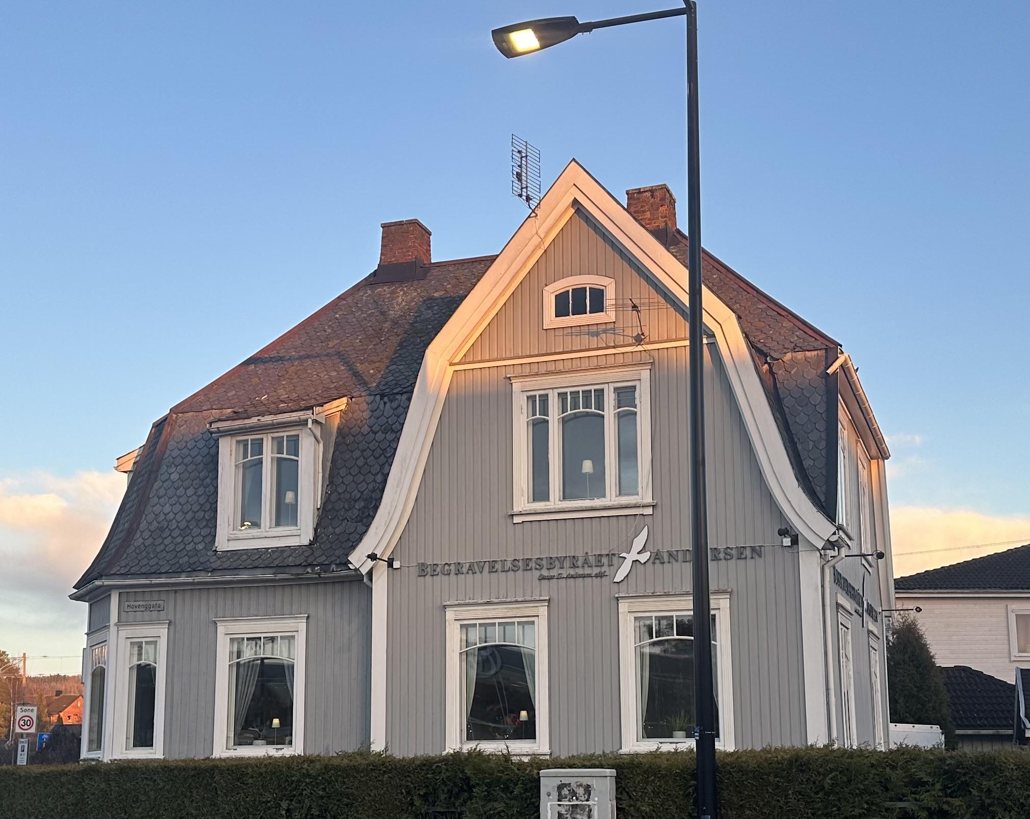 En grå trehus med hvite detaljer og skråtak, beliggende i et boligområde under blå himmel.