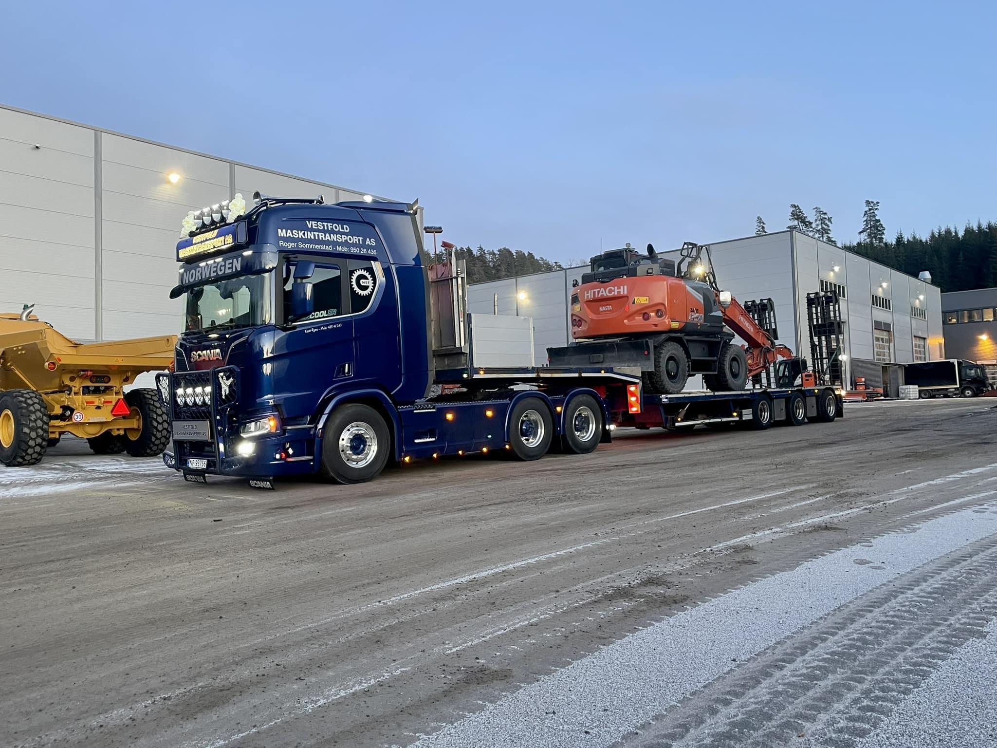 En lastebil med en stor gravemaskin og en annen maskin på tilhenger, parkert ved en lagerbygning.