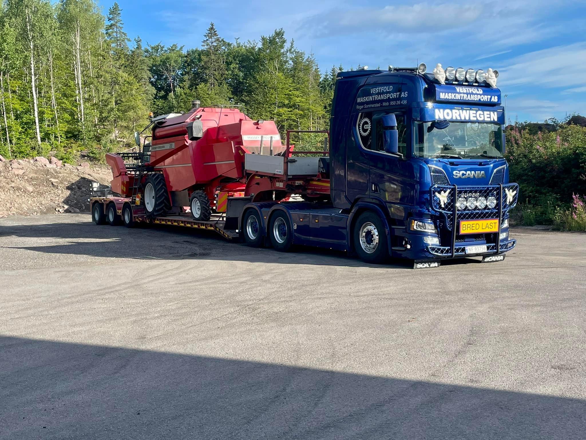 En blå Scania lastebil med tilhenger som frakter en rød maskin, parkert i et skogsområde.