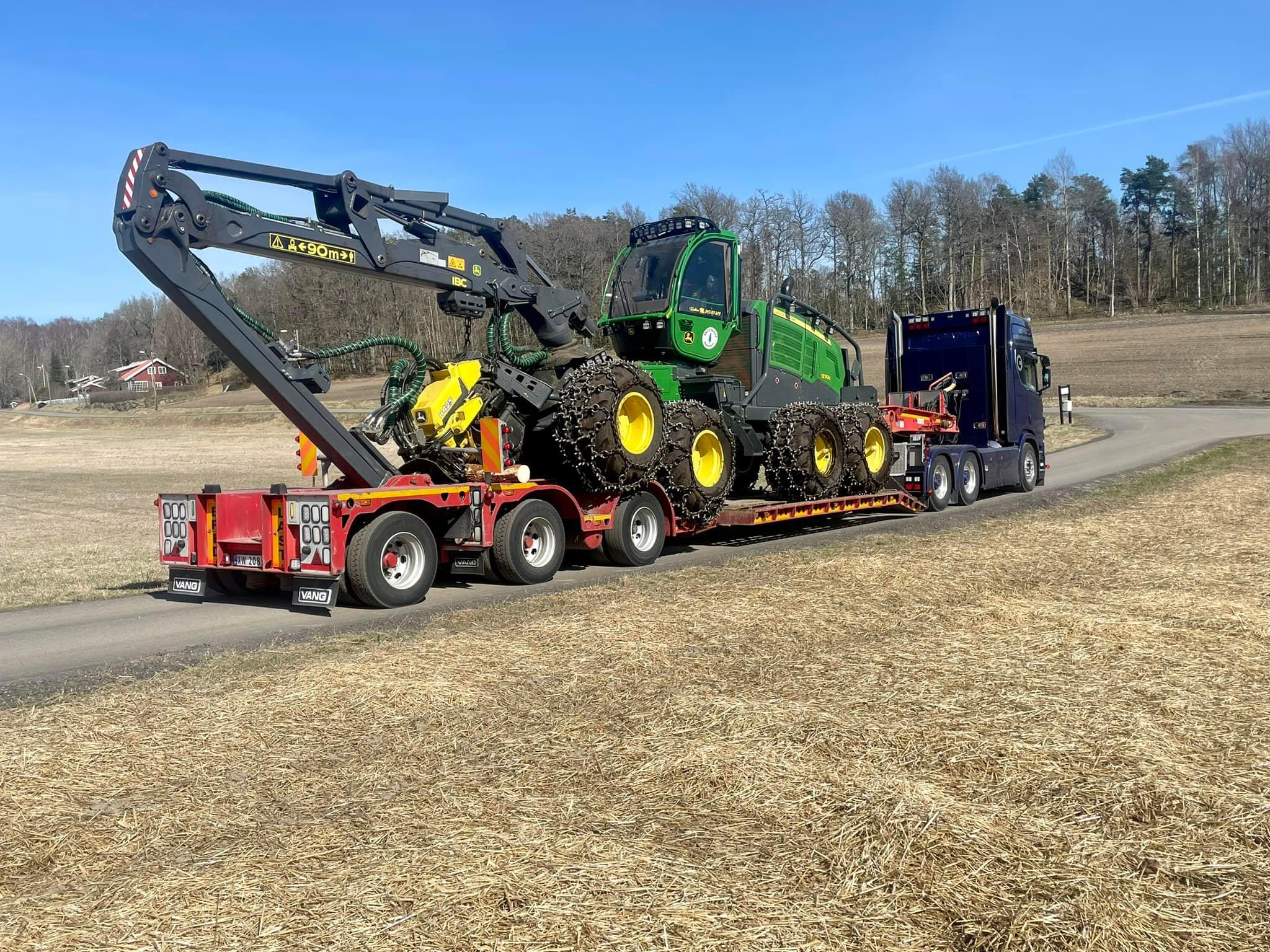 En stor lastebil med en grønn traktor lastet på tilhenger, parkert på en åpen mark.
