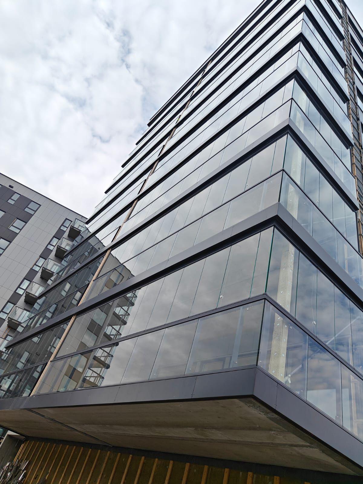 Moderne høyhus med glassfasade og flere etasjer, omgitt av andre bygninger under en skyet himmel.