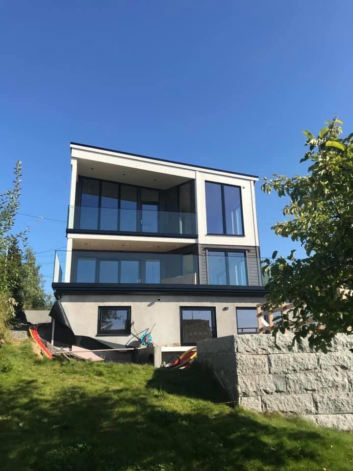 Moderne tre-etasjers hus med store vinduer og balkong, omgitt av grønt gress og trær under en klar blå himmel.