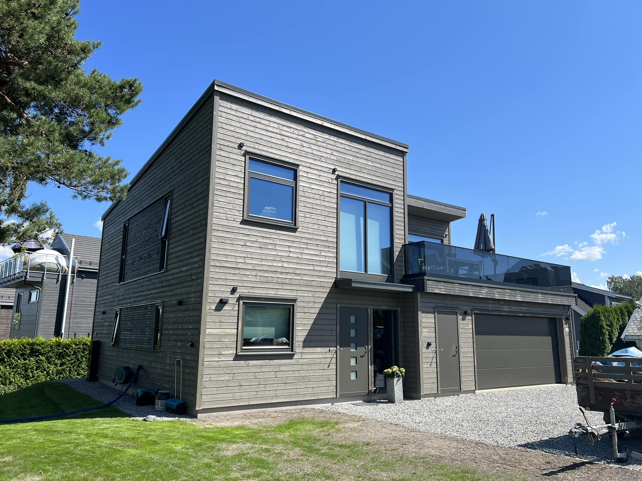 Moderne enebolig med stor terrasse, garasje og grønt område foran, under klar blå himmel.