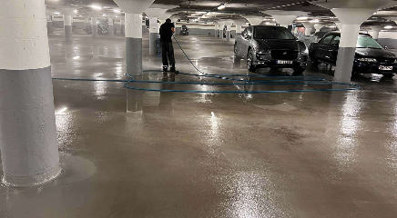 En person vasker gulvet i en parkeringskjeller med en høytrykkspyler, mens biler står parkert.