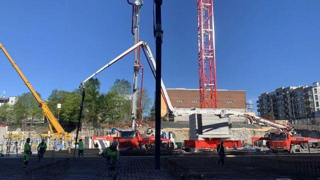 Byggeplass med kraner og arbeidere under solfylt himmel, som jobber med konstruksjon av en bygning.