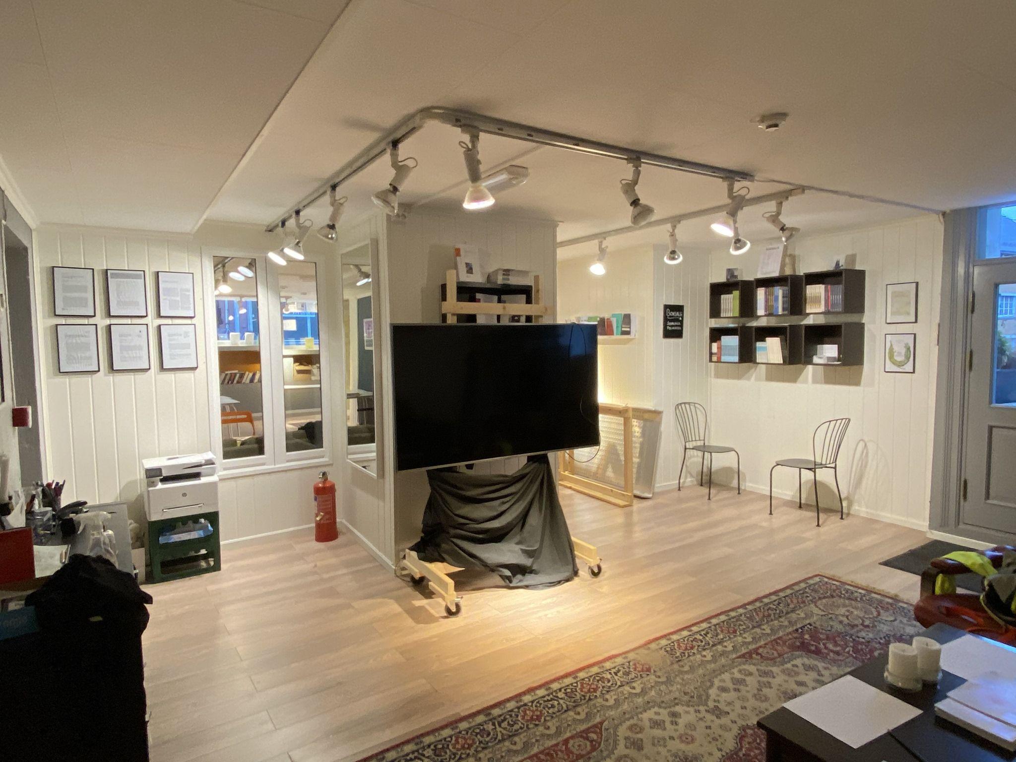 Moderne kontorinnredning med stor TV, hyller, stoler og lyssetting i et lyst rom.