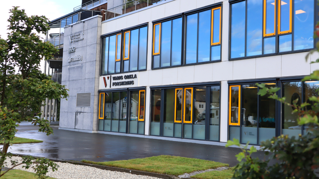 Moderne kontorbygg med store vinduer og gule rammer, beliggende i et grønt område.