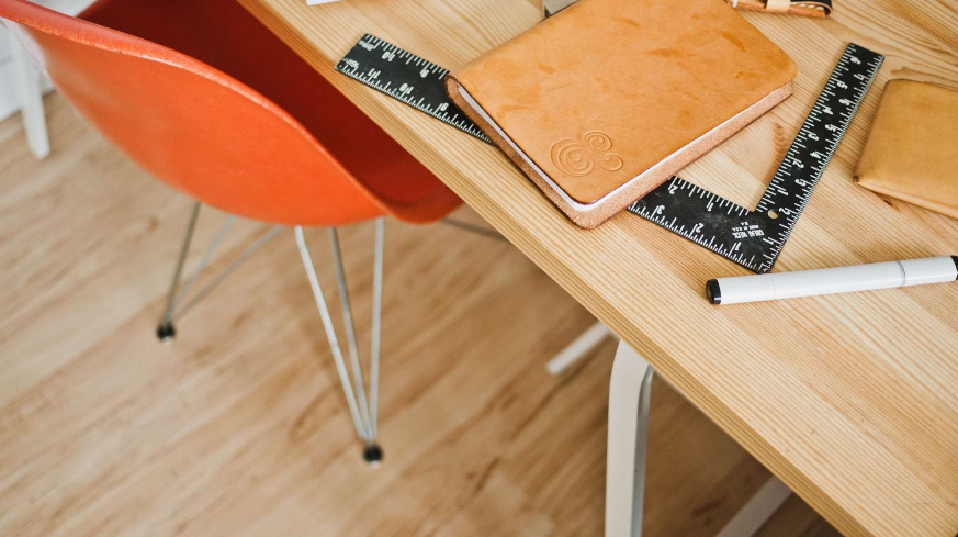 Et skrivebord med en oransje stol, notatbok, linjal og en hvit tusj på treoverflaten.