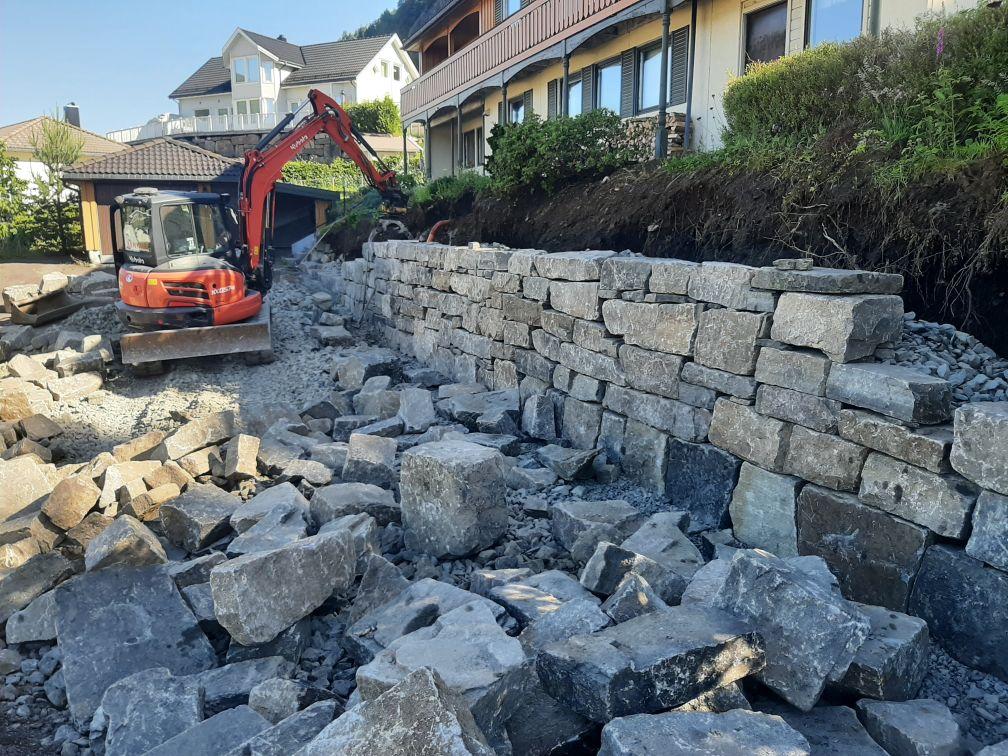 Gravemaskin arbeider med å bygge en steinmur ved en bolig, med steiner spredt rundt området.