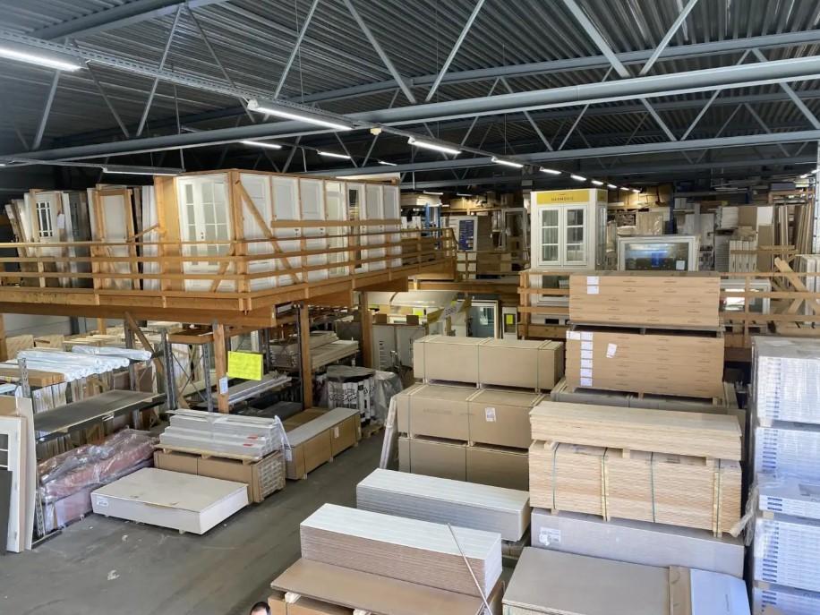 Lager med byggevareprodukter, inkludert skap, plater og materialer stablet i hyller og på gulvet.
