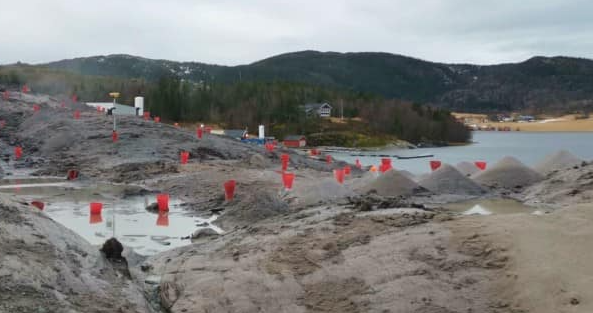 Byggeplass med sandhauger og røde markører ved vannkanten, omgitt av fjell og trær.