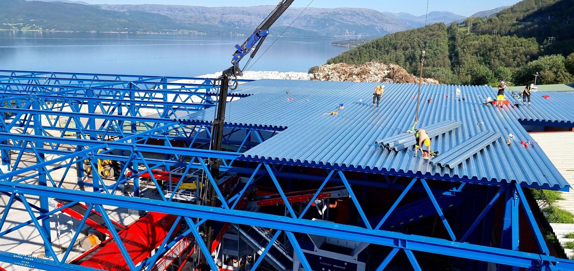 Bygging av et stort tak med blå metallplater, arbeidere i sikkerhetsutstyr og en kran i bakgrunnen ved en innsjø.