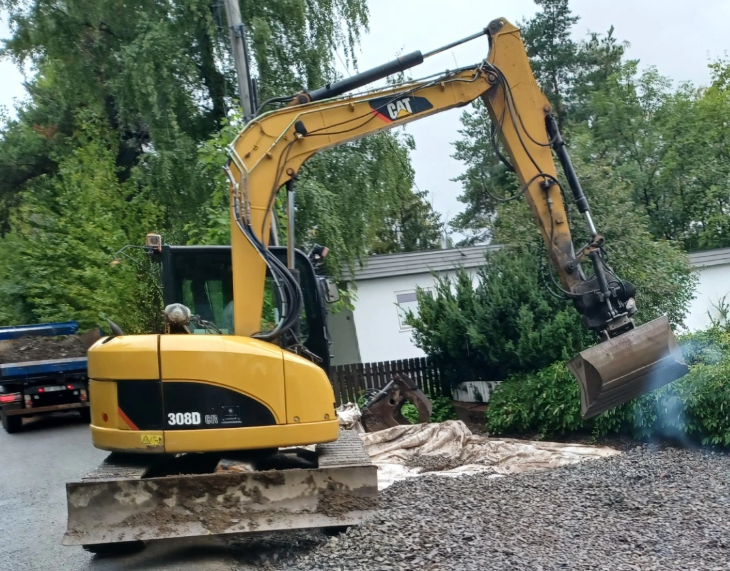 Gravemaskin i arbeid på en hage, med skuffen hevet og klar til å grave. Grøntområde i bakgrunnen.